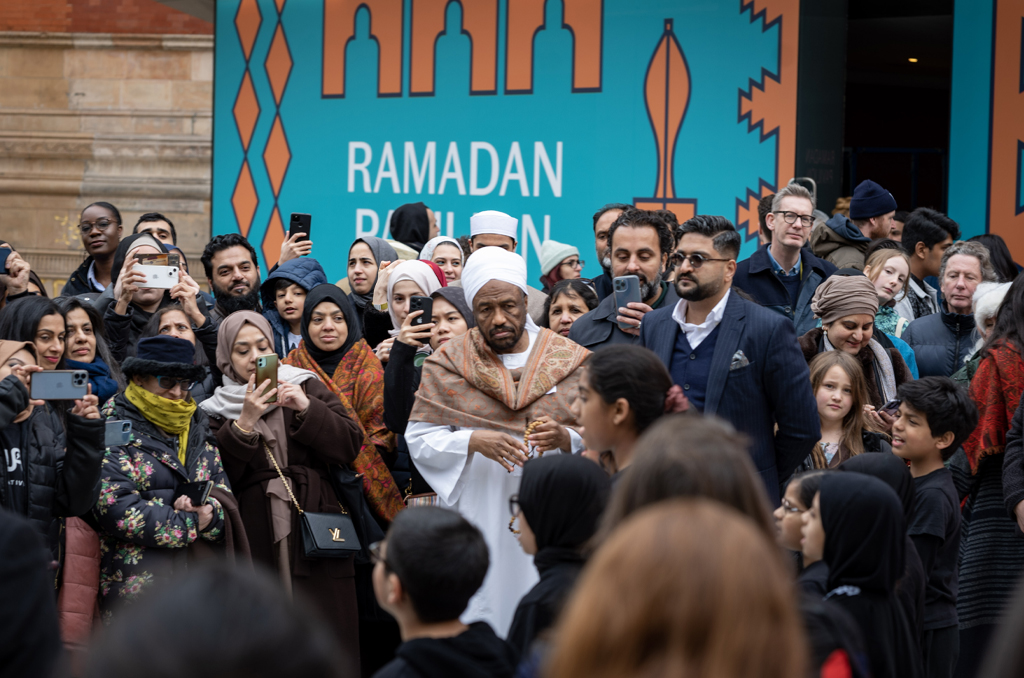 A lot of people taking pictures of the children singing something with blue logo of Ramadan on the back.